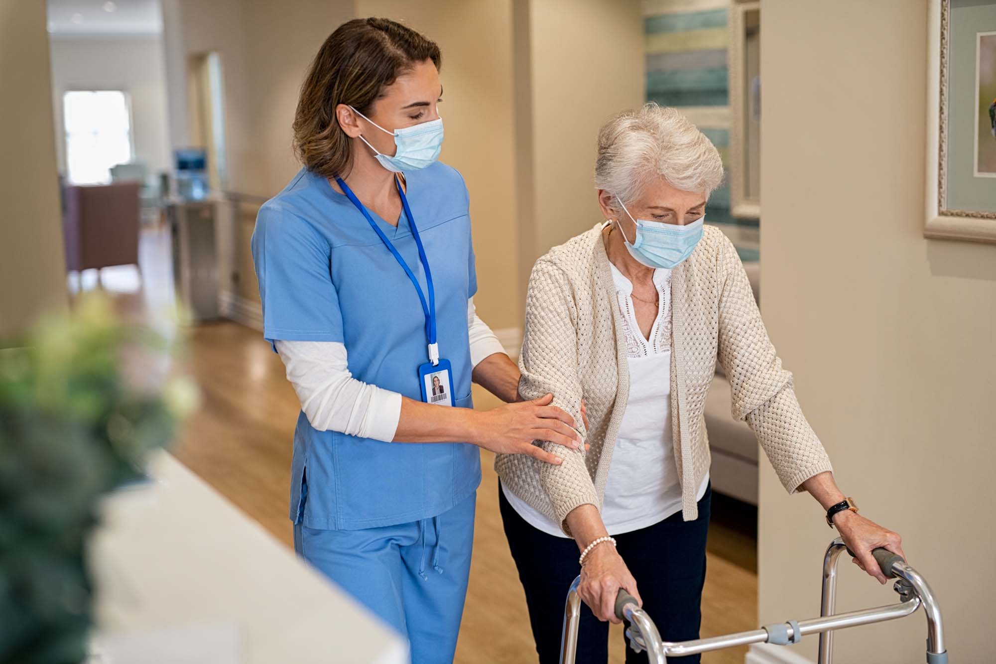 Nurse with face mask helping senior woman to walk around the nursing home with walker.